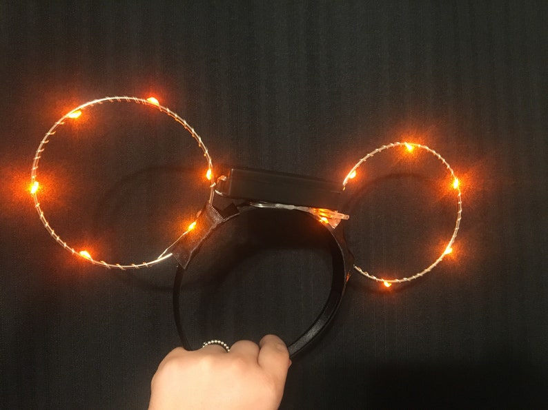 Puede incluir: Una diadema negra con dos c&iacute;rculos de luz naranja unidos a ella. Los c&iacute;rculos est&aacute;n hechos de alambre y tienen peque&ntilde;as luces naranjas incrustadas.