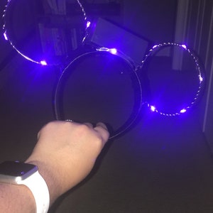 Puede incluir: Un par de orejas de Mickey Mouse negras con luces LED moradas. Las orejas est&aacute;n en la mano de una persona.