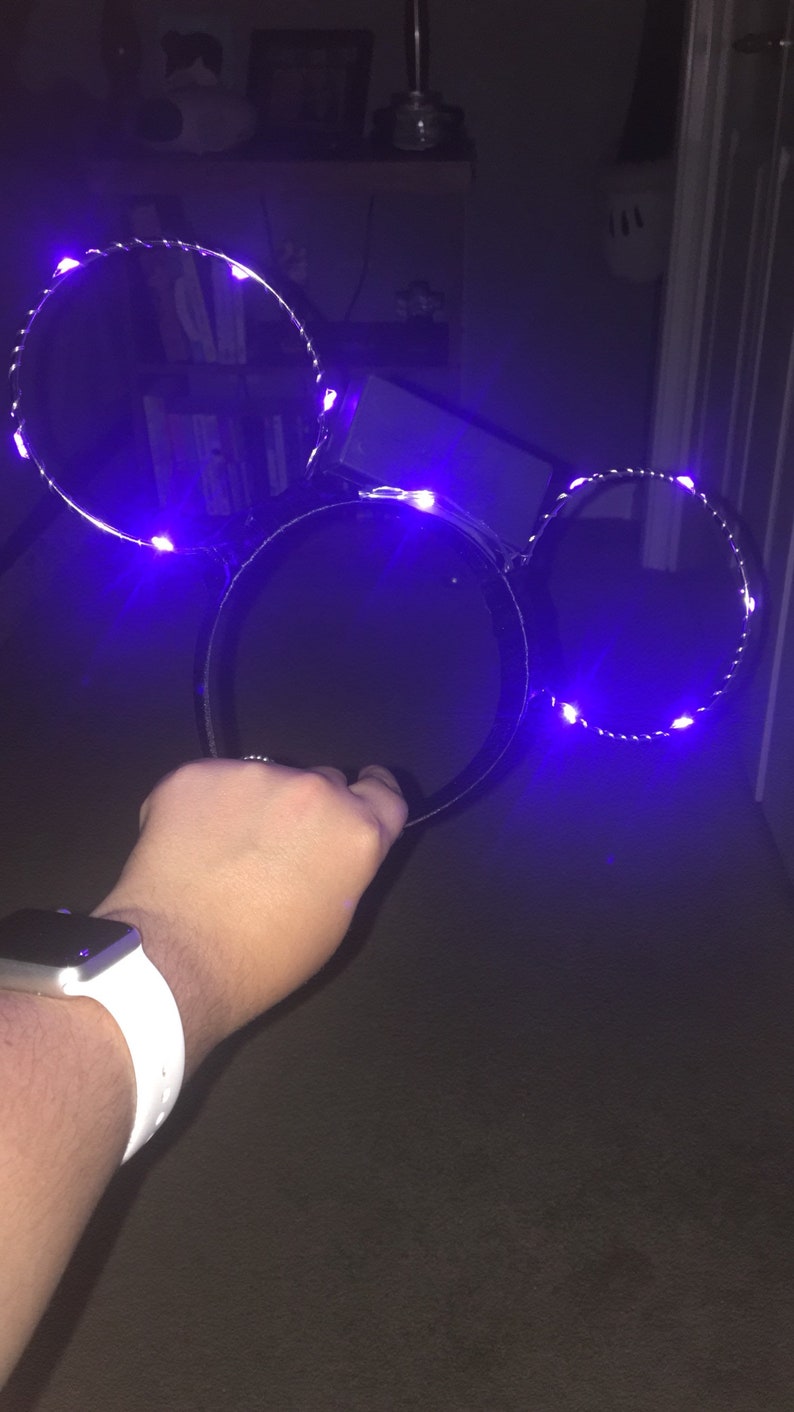 Puede incluir: Un par de orejas de Mickey Mouse negras con luces LED moradas. Las orejas est&aacute;n en la mano de una persona.