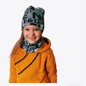 Peut inclure: Une jeune personne portant un bonnet gris et noir à motifs avec le texte "SPORT" et "E&D" dessus. Elle porte également un sweat-shirt orange vif avec un cordon de serrage noir.