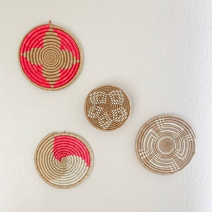 Puede incluir: Cuatro piezas redondas de decoración de pared tejidas en color paja natural con detalles en rosa y blanco. Las piezas están dispuestas en un patrón escalonado en una pared beige claro.