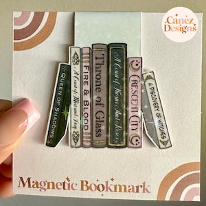 May include: A magnetic bookmark featuring a stack of fantasy book spines. Titles include "Queen of Shadows," "Fire & Blood," and "Throne of Glass." The bookmark has a light pink background with a rainbow design and the text "Magnetic Bookmark."