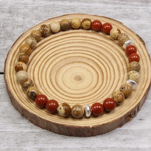 May include: A brown and beige gemstone bracelet with red gemstone accents and silver spacers. The bracelet is on a wooden surface.