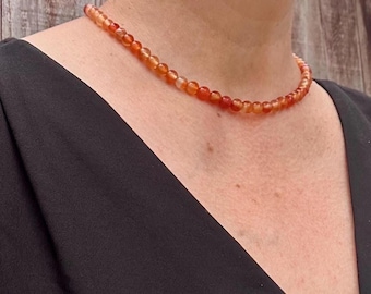 May include: A single-strand necklace composed of small, round, polished carnelian beads in varying shades of translucent orange and deep red. The beads are uniform in size and strung together to form a simple, circular choker-style necklace.