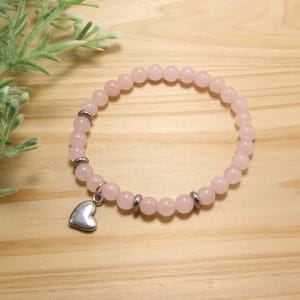 May include: A stretch bracelet composed of round, pale pink beads interspersed with small, polished silver-toned metal spacers. A smooth, silver-toned metal heart-shaped charm hangs from the bracelet, which is arranged in a circular shape.