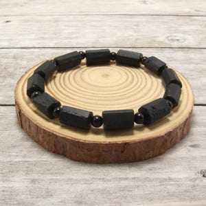 Peut inclure: Un bracelet composé de perles cylindriques noires alternées avec de plus petites perles sphériques noires disposées en cercle. Les perles présentent une finition mate et texturée, assemblées pour former une boucle continue de composants sombres.
