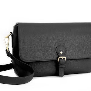 May include: Black crossbody bag with a textured finish. The bag features a flap closure with a gold-colored buckle and a matching adjustable shoulder strap. The bag is designed with a rectangular shape.