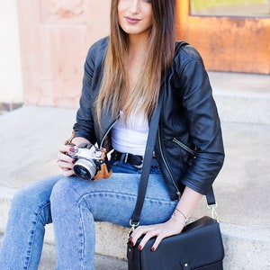 May include: A black leather jacket, white top, and blue jeans. A black crossbody bag with a gold buckle and a vintage camera with a brown leather case. The person is sitting on concrete steps.