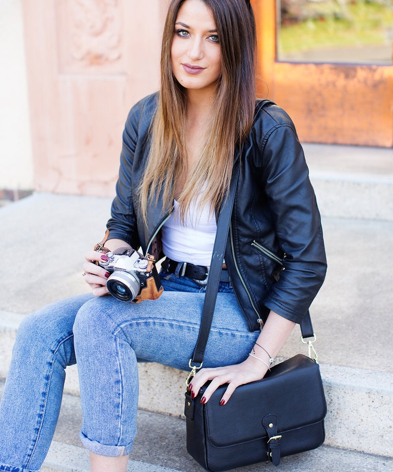 May include: A black leather jacket, white top, and blue jeans. A black crossbody bag with a gold buckle and a vintage camera with a brown leather case. The person is sitting on concrete steps.