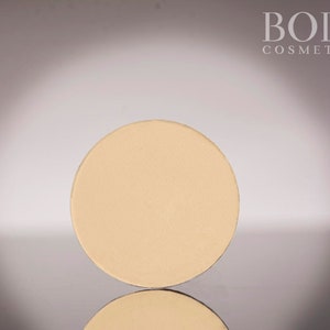 May include: A round, beige-coloured pressed powder compact. The compact is on a white surface with the text "BODI COSMETICS" in black letters at the top of the image.