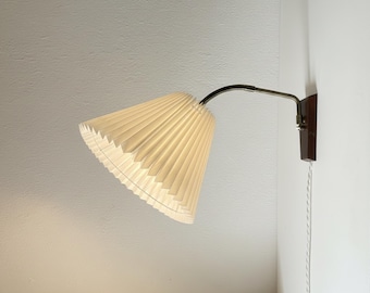 May include: A wall-mounted lamp featuring a white, pleated, cone-shaped shade attached to a curved, metallic arm. The arm connects to a dark brown rectangular wooden base mounted on the wall, with a white electrical cord hanging downward.
