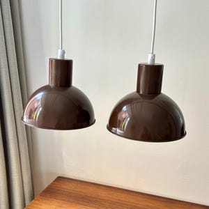 Może przedstawiać: Dwie brązowe lampy wiszące z błyszczącym wykończeniem wiszą na białych przewodach. Lampy mają cylindryczną górę i zaokrąglony, miseczkowaty klosz. Tło obejmuje drewnianą komodę i ścianę w neutralnym kolorze.