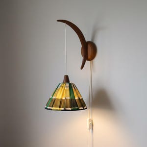 Può includere: Una lampada da parete con un braccio in legno curvo e un paralume conico con motivo a quadri giallo, verde e nero. L'apparecchio include un cavo di alimentazione bianco con interruttore, che scende dal supporto a parete in legno.