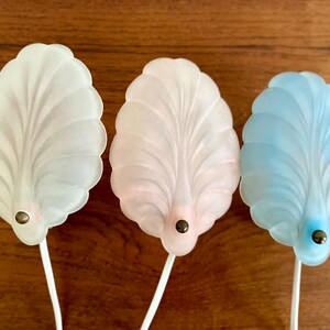 Könnte beinhalten: Drei dekorative Wandleuchten in Form einer Muschel. Die Leuchten sind aus mattiertem Glas und in Weiß, Rosa und Blau erhältlich.