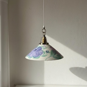 Könnte beinhalten: Eine konische Hängelampe mit einem cremefarbenen Keramikschirm, der handbemalt mit violetten und blaugrünen Blumenmotiven ist. Die Leuchte verfügt über eine messingfarbene Metallfassung und hängt an einem dünnen weißen Stromkabel.