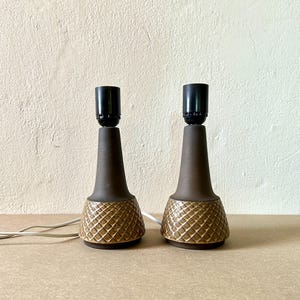 Może przedstawiać: Dwie brązowe ceramiczne lampy stołowe z czarnymi oprawkami. Lampy mają teksturowaną podstawę w romby i gładki, zwężający się korpus. Lampy są połączone z białym przewodem elektrycznym.