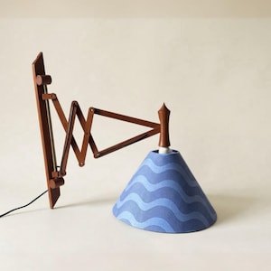 May include: A wall-mounted lamp featuring a brown wooden accordion-style extendable arm and a conical lampshade. The shade displays a repeating pattern of wavy horizontal lines in alternating shades of light and dark blue, with a textured finish.