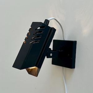 May include: A black wall-mounted lamp with a rectangular shade and a square base. The shade has a geometric design with cut-out details. A white cord extends from the top of the lamp. The lamp is mounted on a white wall.