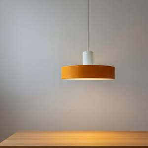 May include: A modern pendant light fixture with a cylindrical white top and a wide, orange-colored, circular shade. The light hangs from a thin white cord above a wooden table, creating a warm, inviting ambiance. The fixture is a simple, minimalist design.