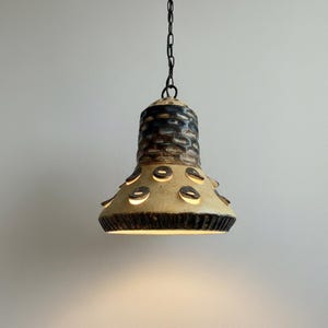 Puede incluir: Una lámpara colgante de cerámica con una pantalla en forma de campana que presenta una sección superior texturizada de color marrón oscuro y una sección inferior crema adornada con acentos circulares en relieve de color marrón oscuro.