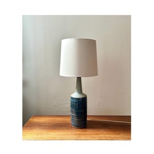 Könnte beinhalten: Eine Keramik-Tischlampe mit einem weißen Stofflampenschirm. Die Lampenbasis ist eine blaue und weiße glasierte Keramikflasche.