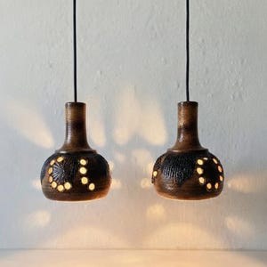 May include: Two brown ceramic pendant lights with a bulbous shape and narrow necks. Each fixture features intricate cut-out patterns that allow light to shine through, creating a decorative glow. The lamps hang from thin black cords.