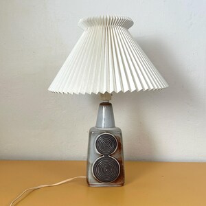 May include: A vintage table lamp with a white pleated lampshade. The lamp base is gray with a brown, circular design. The lamp is plugged into an electrical outlet.