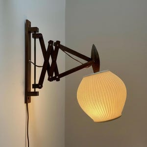 Könnte beinhalten: Eine Wandleuchte mit einem ausziehbaren Scherengitterarm aus dunkelbraunem Holz. Die Lampe hat einen abgerundeten, strukturierten weißen Lampenschirm mit einer gefalteten Oberfläche, der ein warmes Licht ausstrahlt.