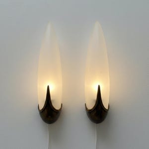 Pode incluir: Um par de arandelas de parede com cúpulas de vidro fosco em formato de gota alongada. Cada cúpula é sustentada por uma base de metal em tom bronze escuro com um design pontiagudo semelhante a uma pétala. As lâmpadas emitem um brilho quente.