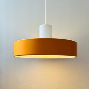 Może przedstawiać: Nowoczesna lampa wisząca z cylindrycznym białym topem i szerokim, okrągłym kloszem w ciepłym pomarańczowym kolorze. Światło zwisa z białego przewodu, oświetlając przestrzeń poniżej. Oprawa ma minimalistyczny design.