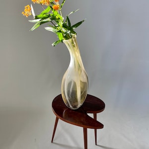 May include: A white glass vase with a unique shape and a brown wooden side table with three legs. The vase is filled with orange flowers.