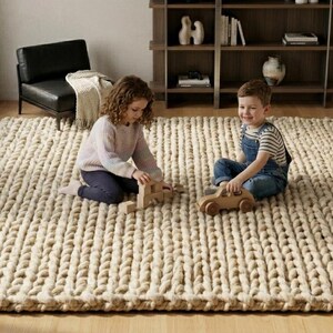 Puede incluir: Una alfombra grande, rectangular, tejida a mano, de color blanco roto natural. Dos ni&ntilde;os juegan en la alfombra con juguetes de madera. La gruesa construcci&oacute;n de punto grueso de la alfombra a&ntilde;ade una textura acogedora a la habitaci&oacute;n.