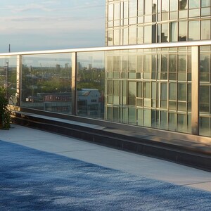 May include: A modern rooftop scene featuring a large, textured blue rug. A glass railing and a high-rise building with reflective windows are in the background. The sky is a clear blue.