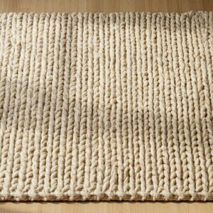 Puede incluir: Una alfombra rectangular de punto grueso de color crema. La alfombra presenta un patr&oacute;n de costillas verticales, hecho de hilo grueso y suave. Se coloca sobre un suelo de madera clara. Un acogedor elemento de decoraci&oacute;n del hogar, aprox. 150 cm x 200 cm.