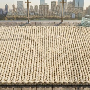 Puede incluir: Una alfombra grande, rectangular, de punto grueso en color beige natural. La alfombra est&aacute; hecha de hilo grueso y tiene una superficie texturizada. Se coloca sobre una terraza de madera con un horizonte urbano al fondo.