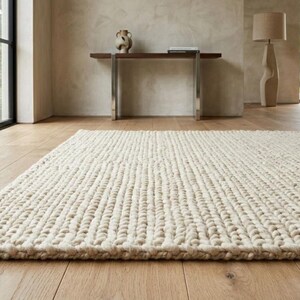 Puede incluir: Una alfombra grande, rectangular, de color crema con un tejido grueso y texturizado. La alfombra est&aacute; colocada sobre un suelo de madera clara en una habitaci&oacute;n con grandes ventanales y una est&eacute;tica minimalista. Las dimensiones de la alfombra no son visibles.
