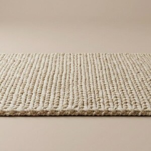 Puede incluir: Una alfombra rectangular de color beige con una textura gruesa y tejida. La alfombra est&aacute; hecha de hebras verticales gruesas, creando una superficie texturizada. La alfombra se coloca sobre un fondo neutro, resaltando su dise&ntilde;o simple y color natural.