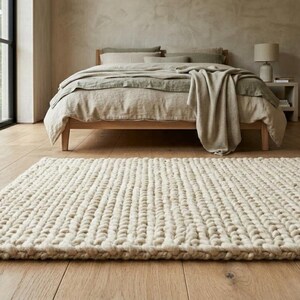 Puede incluir: Una alfombra rectangular de punto grueso de color crema se encuentra sobre un suelo de madera clara frente a una cama. La cama tiene un marco de madera y est&aacute; cubierta con ropa de cama de tonos neutros. Se ve una ventana grande con marco negro.