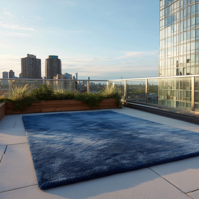 May include: A large, rectangular blue rug on a rooftop patio. The rug has a textured appearance, with varying shades of blue. The background features a cityscape with tall buildings and a clear blue sky. The rug is a decorative element for outdoor spaces.