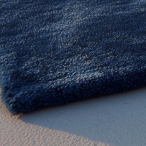 May include: A close-up of a section of a navy blue rug with a textured surface. The rug's corner is visible, showing the thickness and plushness of the material. The rug is placed on a light-colored, textured surface.