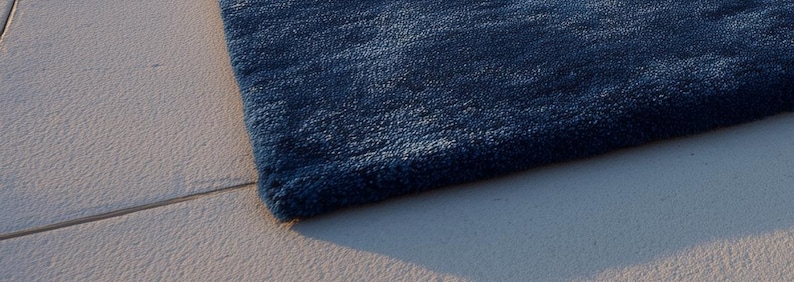 May include: A close-up of a section of a navy blue rug with a textured surface. The rug's corner is visible, showing the thickness and plushness of the material. The rug is placed on a light-colored, textured surface.