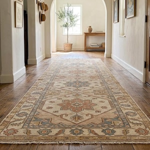 May include: A long, rectangular rug with an intricate geometric pattern in shades of beige, peach, and blue. The rug is placed on a dark wood floor in a hallway, with white walls and a wooden door visible in the background.