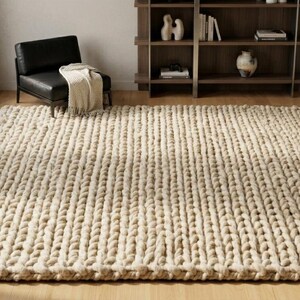 Puede incluir: Una alfombra grande, rectangular, de punto grueso en color beige neutro. La alfombra est&aacute; colocada sobre un suelo de madera, con una silla de cuero negro y una estanter&iacute;a de madera en el fondo. La alfombra mide unos 244 x 305 cm.