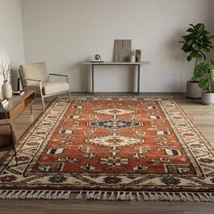 Peut inclure: Un grand tapis rectangulaire au design traditionnel. Le tapis présente un fond de couleur rouille avec un motif géométrique central bleu, blanc et crème. La bordure présente un motif répétitif et le tapis a une frange sur les bords.