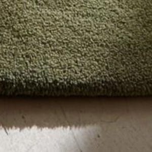 May include: Close-up of a green, textured rug. The rug has a thick pile and a soft appearance. The color is a muted green, and the texture appears plush. The rug's edge is visible, showing the depth of the fibers.