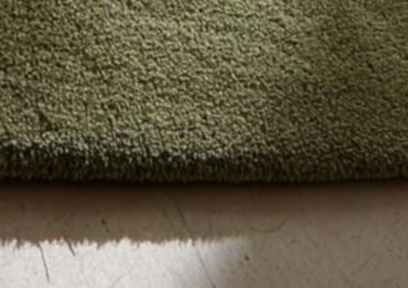 May include: Close-up of a green, textured rug. The rug has a thick pile and a soft appearance. The color is a muted green, and the texture appears plush. The rug's edge is visible, showing the depth of the fibers.