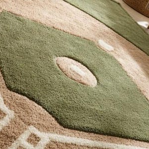 May include: Close-up of a large area rug with a geometric design. The rug features a diamond shape in olive green with a beige oval in the center. The background is beige with white geometric patterns.