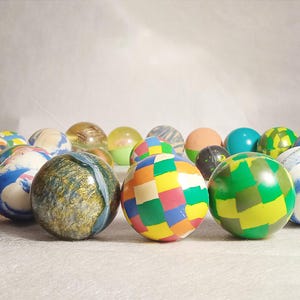 May include: A collection of colorful bouncy balls in various patterns and colors, including green, blue, orange, and yellow. The balls are arranged in a semi-circle, showcasing their diverse designs.