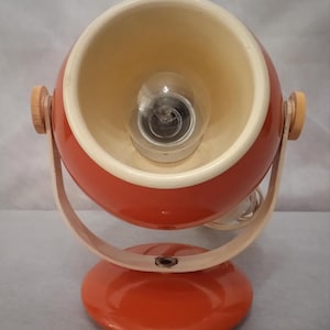 Könnte beinhalten: Retro-Schreibtischlampe in Orange und Creme. Die Lampe hat einen kugelförmigen, orangefarbenen Schirm mit einer Glühbirne, der von einem gebogenen Holzrahmen getragen wird. Der Sockel ist rund und orange, mit orangefarbenen Knöpfen an den Seiten.