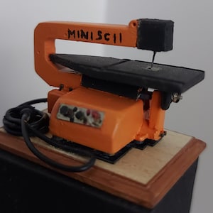 Peut inclure: Une scie sauteuse miniature orange et noire avec le texte "MINISCII" sur le bras, posée sur une base en bois.